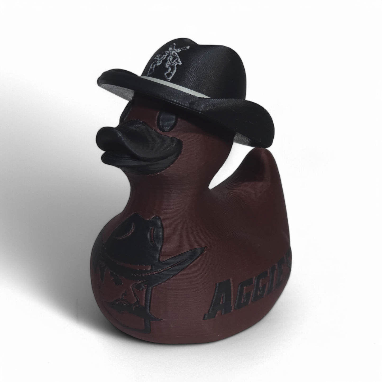 A dark brown 3D printed PLA duck styled as a cowboy, wearing a black hat with a silver emblem and featuring a black cowboy hat design and the word 'AGGIE' on its side, set against a plain white background.