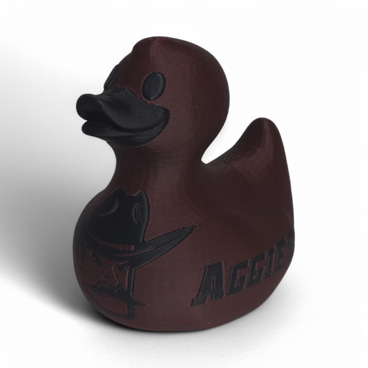 A dark brown 3D Printed PLA duck with a matte finish, featuring a black embossed cowboy hat logo and the word 'AGGIES' on the side, set against a plain white background.