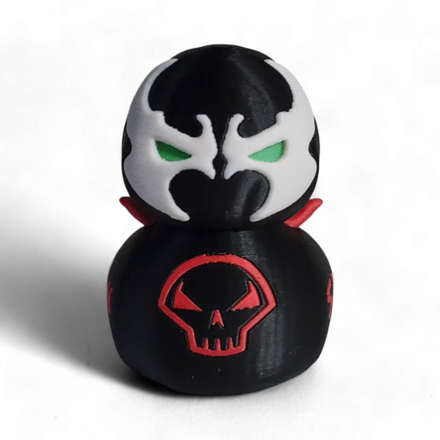 A small black figurine shaped like a duck or duck-head character, with a white mask design, green eyes, red lips, a red skull emblem on the front, and the word 'SPAWN' in red on the side, photographed on a white background.