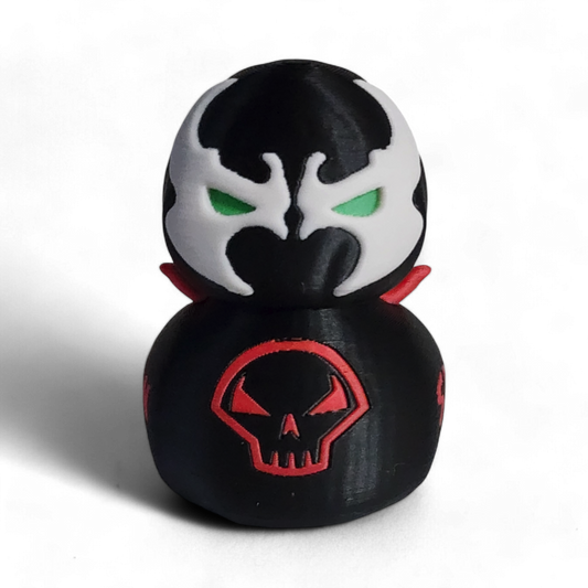 A small black figurine shaped like a duck or duck-head character, with a white mask design, green eyes, red lips, a red skull emblem on the front, and the word 'SPAWN' in red on the side, photographed on a white background.