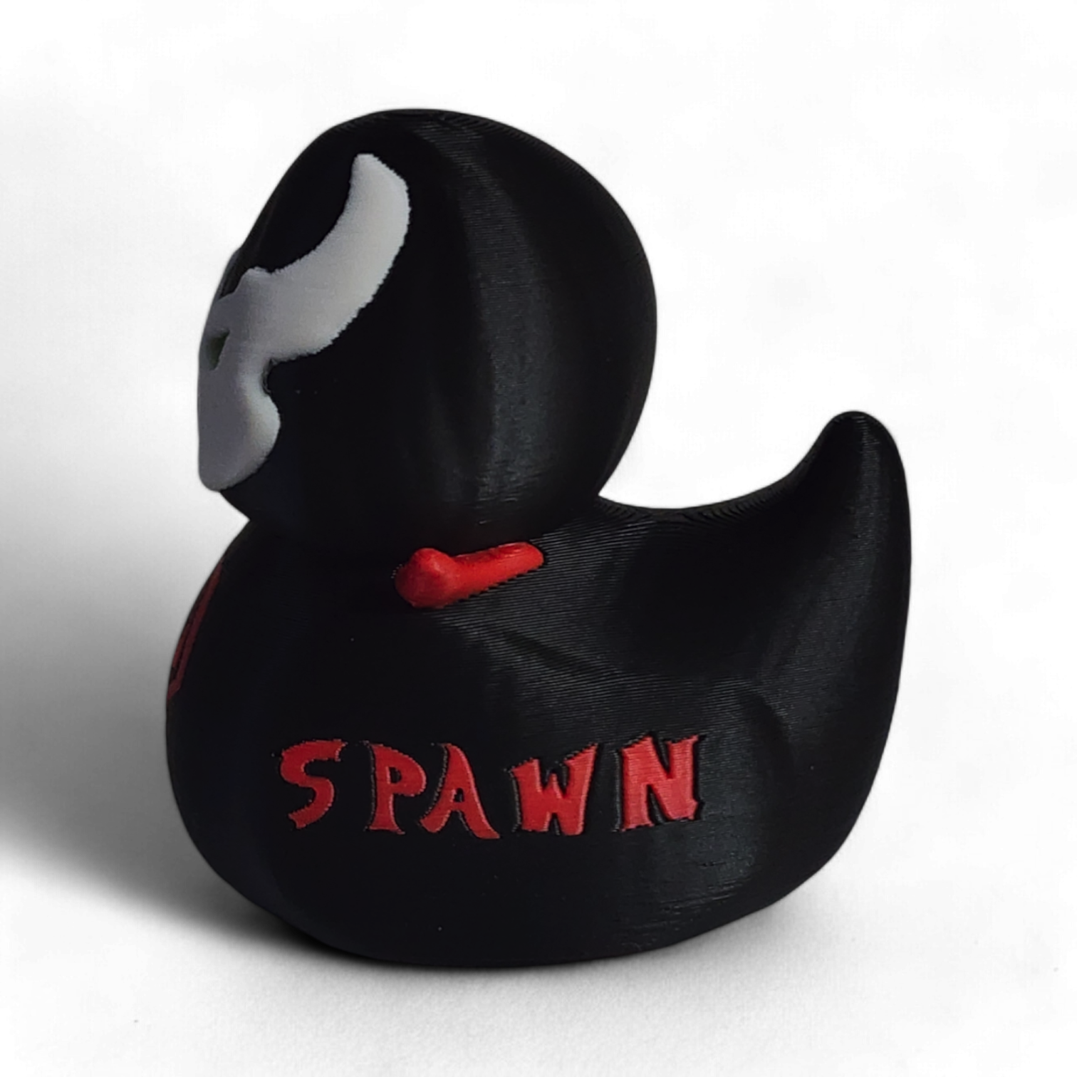 A small black figurine shaped like a duck or duck-head character, with a white mask design, green eyes, red lips, a red skull emblem on the front, and the word 'SPAWN' in red on the side, photographed on a white background.