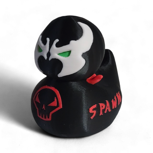 A small black figurine shaped like a duck or duck-head character, with a white mask design, green eyes, red lips, a red skull emblem on the front, and the word 'SPAWN' in red on the side, photographed on a white background.
