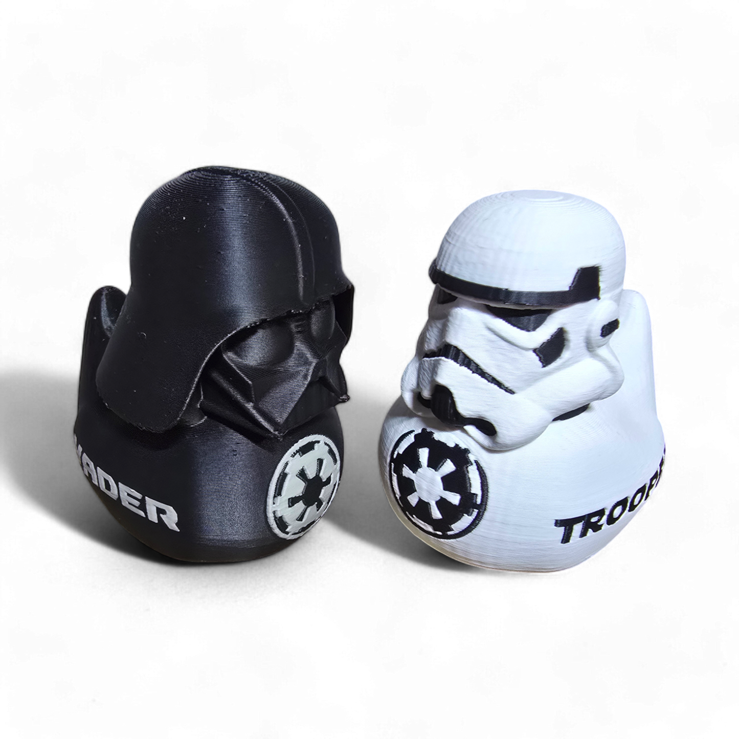 Two small Star Wars character-shaped items on a plain white background: a black Darth Vader figure with a white Imperial crest and partial white 'RADER' text, and a white Stormtrooper figure with a black crest and partial black 'TROOP' text.