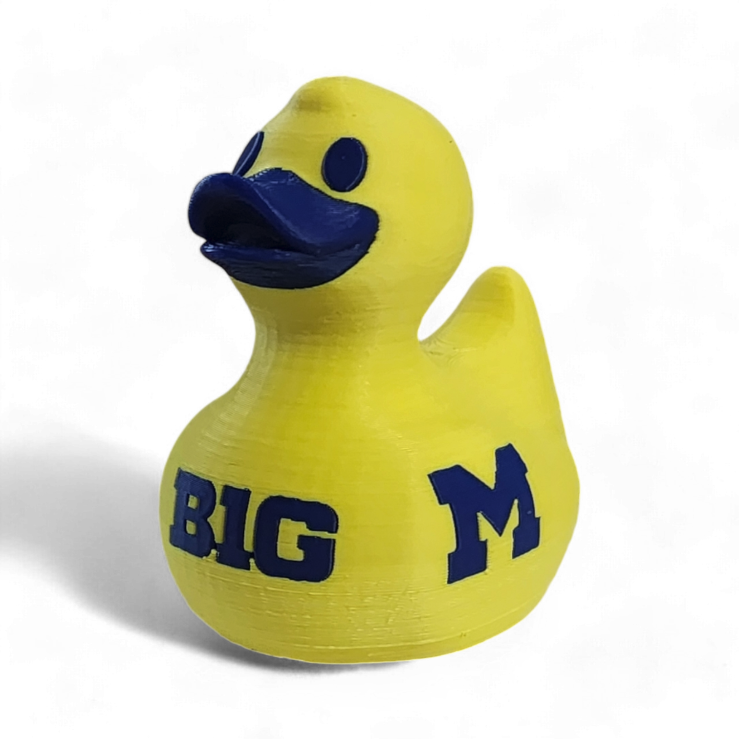 A yellow 3D printed duck with a dark blue beak and eyes, featuring the text 'BIG' and a large letter 'M' printed in dark blue on the side, set against a plain white background.