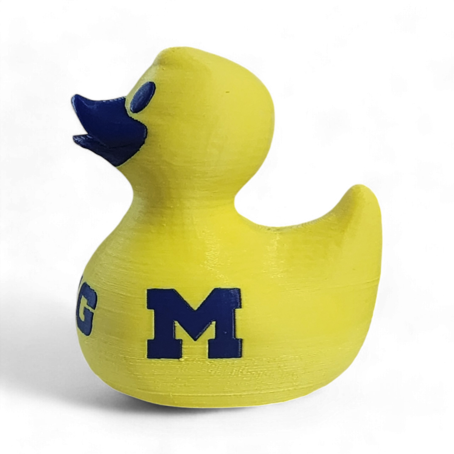 A yellow 3D printed duck toy with a dark blue beak and a large navy blue letter 'M' on the side, representing the Michigan State University logo, on a white background.