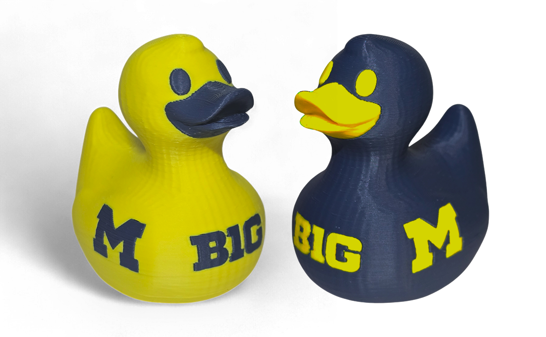 Two 3D printed duck toys with a 3D-printed look, one yellow and one navy blue, both featuring the letters 'M' and 'BIG' in the opposite color. They are shown facing each other on a white background.