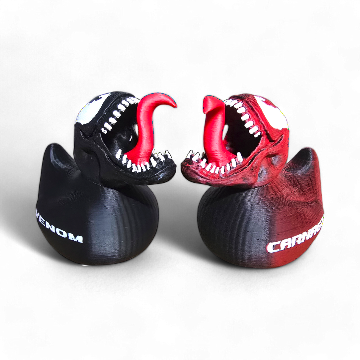 Two 3D-printed shark head-shaped standees or figurines facing each other, one black with white and red details labeled VENOM, and one red with white and black details labeled CARABAS, on a light gray background.