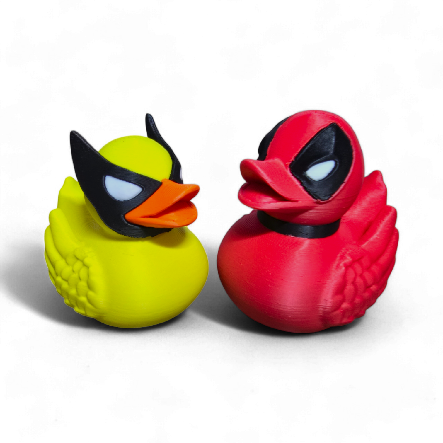 Two 3D-printed rubber duck toys styled as Marvel characters, one in yellow with a black and orange mask resembling Wolverine, and the other in red with a black and white mask resembling Deadpool, set against a plain white background.